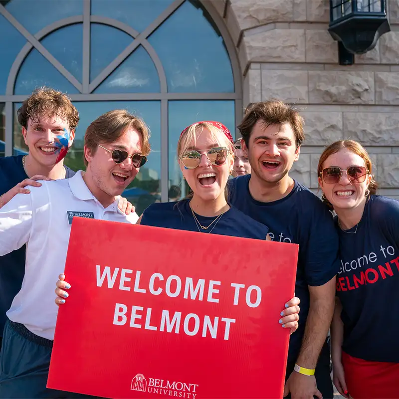 A group of five enthusiastic students standing in front of a stone building, holding a red sign that reads ‘Welcome to Belmont,’ all wearing Belmont University gear.
