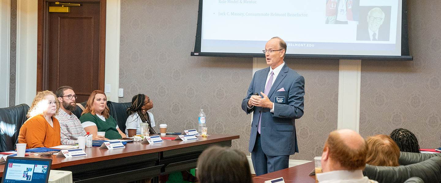 A man in a suit giving a presentation to a group of people seated around a conference table, with a PowerPoint slide projected on the wall behind him.