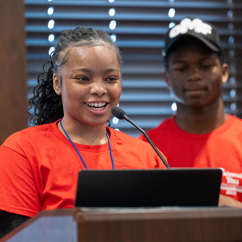 A young woman in a red T-shirt speaks confidently at a podium with a microphone, while a young man in a matching red T-shirt and a black cap stands behind her, listening attentively.