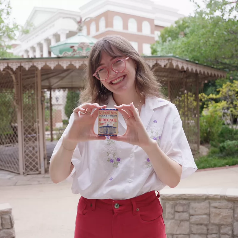 Emily Henegar holding a cookie with phrase "Belmont Boys make their Birdcage debut" outdoors. She wears a white shirt, red glasses and red pants, standing in front of a gazebo that Belmont students refer to as the "the Birdcage."