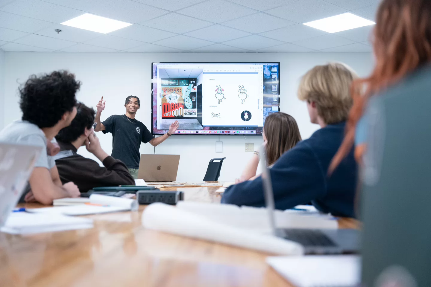 A person presents to a group of students in a classroom, pointing at a screen displaying colorful graphics. The atmosphere is engaging and attentive.