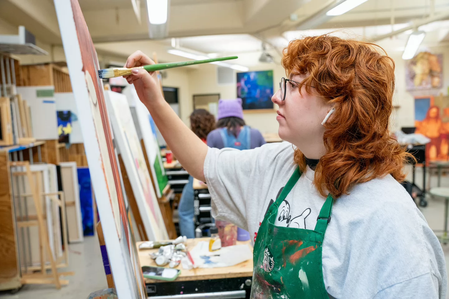 A person with curly red hair paints on a canvas in a studio, wearing glasses and a green apron. The setting is bright and cluttered with art supplies.