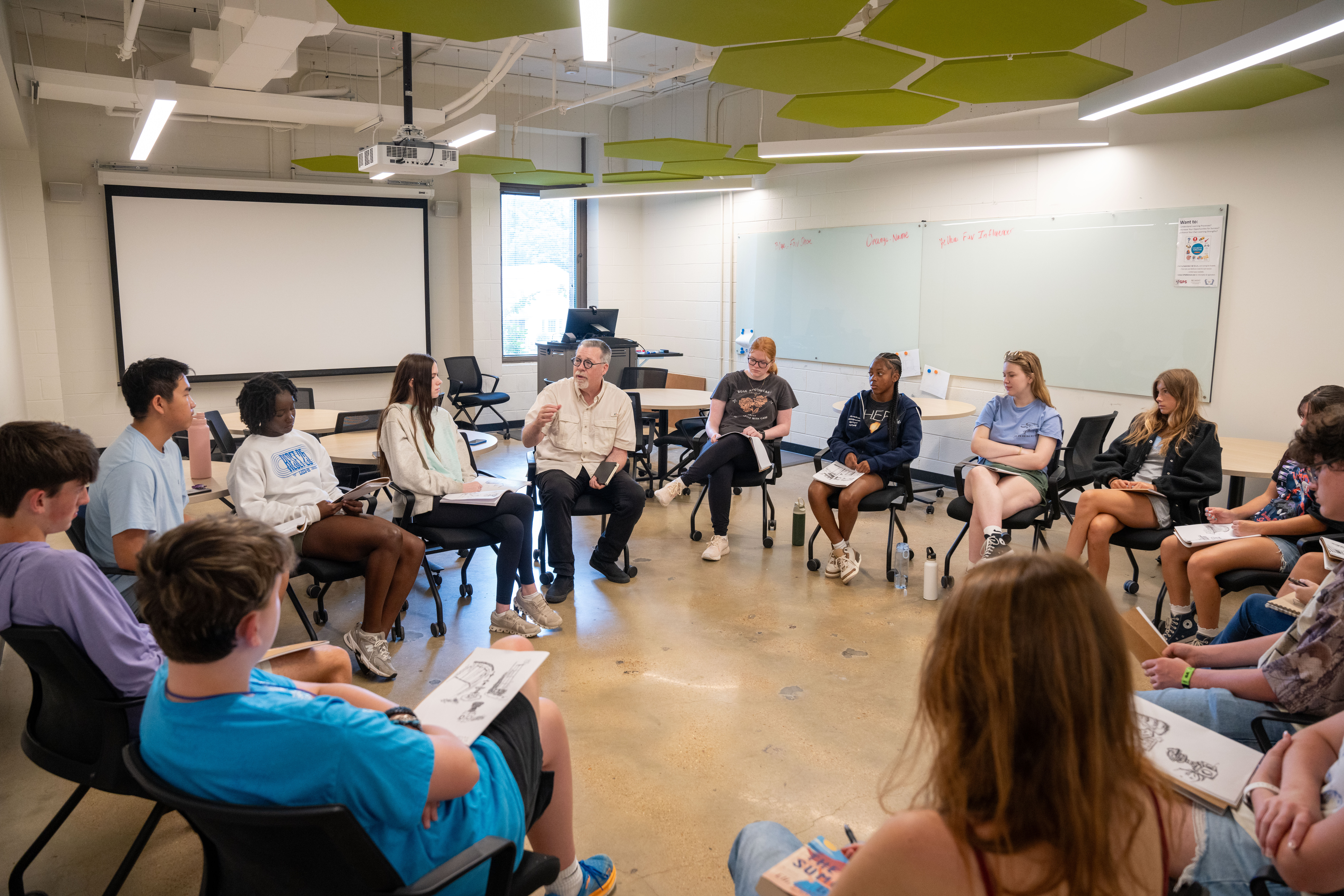 Teen summer program students listen to teacher lecture, while seating in a circle. Students all have sketchbooks with them. 