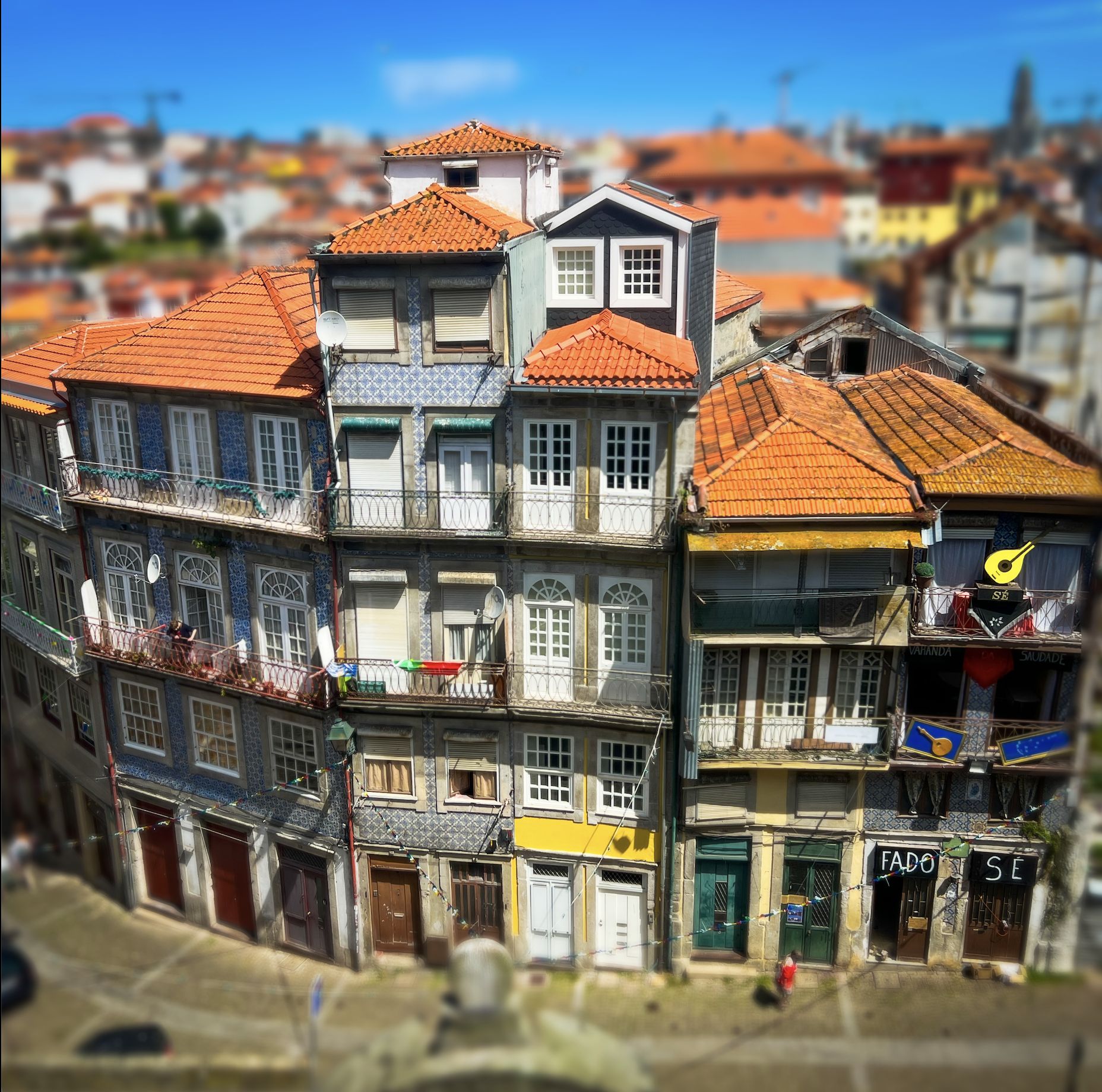 Colorful European-style buildings with red-tiled roofs and balconies displaying clothes and flags, lining a cobblestone street with pedestrians.