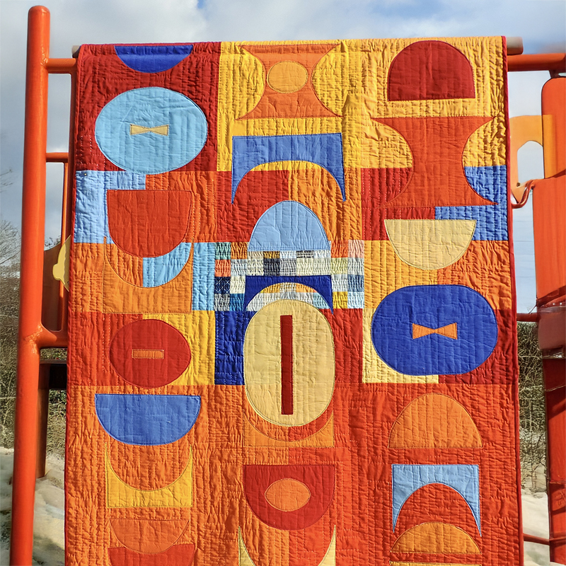 Image of a blue and orange quilt hanging over playground equipment to display geometric quilt pattern
