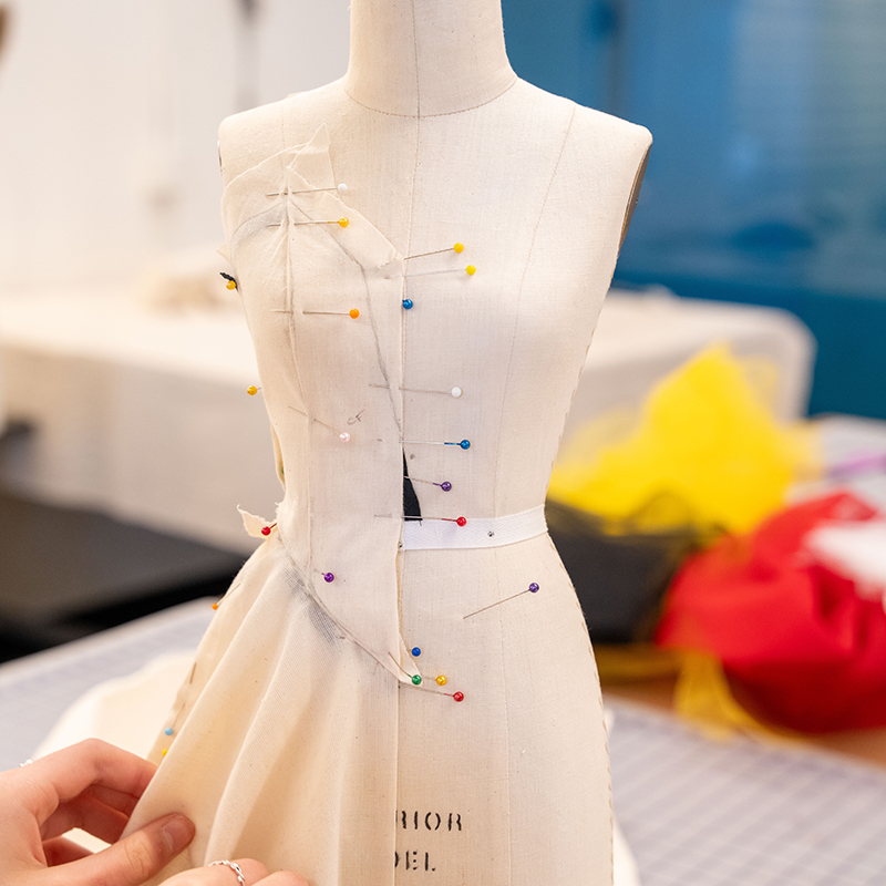 A dress form with white straight pins holding on a piece of white muslin draped to make a dress. 