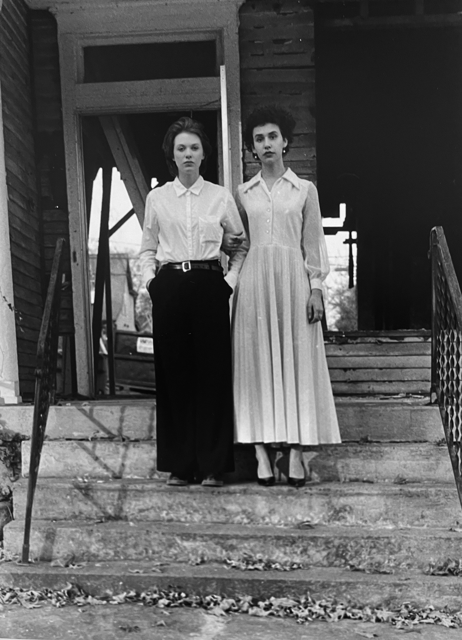 2 women wearing long skirts on the porch of an old house in black and white